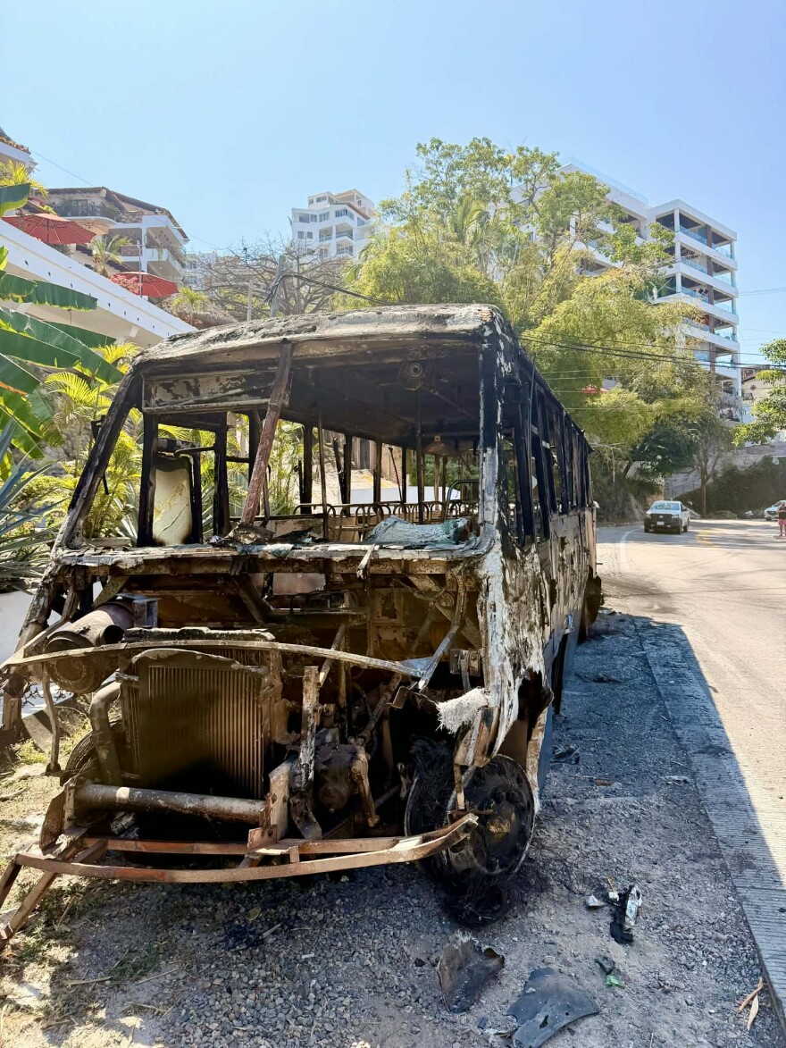The burnt and rusted metal shell of a bus that was previously lit on fire sits on the side of a paved road. 