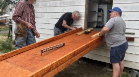 Three men are gathered around the back door of a house. They are working on attaching a wooden wheelchair ramp to the house.