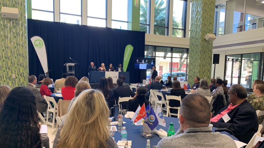 panel of people speaking at a press conference, people seated at tables listening to them.