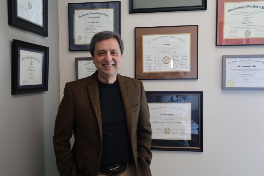 Dr. Ali Hammad stands in front of a wall in his office. The wall has framed diplomas and certificates.