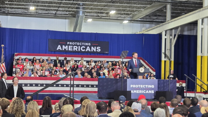 Vice President JD Vance speaks at a podium before a large audience seated in an industrial venue. Red, white, and blue decorations surround the stage, with signs reading “Protecting Americans.” A teleprompter stands beside Vance, and security personnel and supporters are visible in the scene.