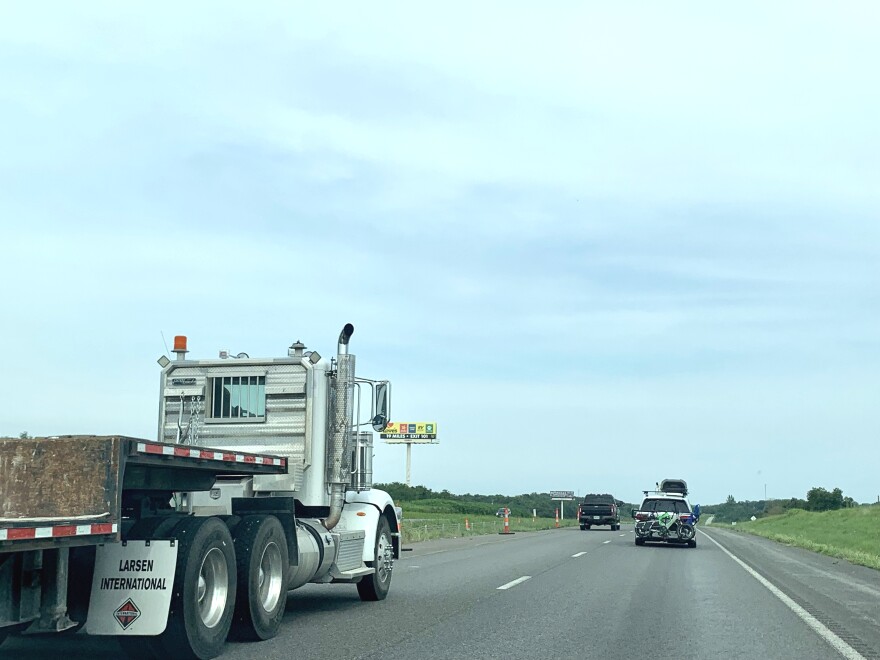 A view of I-70 from the front seat of a car. A semi truck drives by on the left hand side of the frame. A van with bikes attached to the back is in the right lane. Traffic cones are visible in the distance. 
