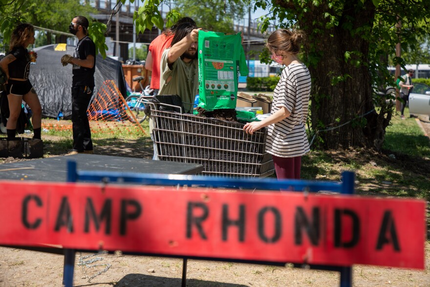 Foreground: A red sign with black letters reads "Camp Rhonda." Background: Two volunteers pour a green bag of mulch into a shopping cart lined with fabric.