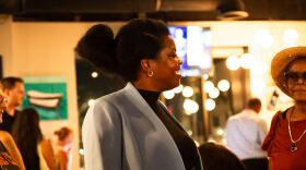 A Black woman in a black turtleneck and gray or light blue blazer stands in profile in a photo at an indoor election watch party. 
