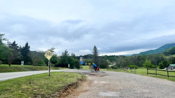 A bicyclist is riding on gravel next to a road. A yellow bicycle sign is on the right side of the road. A mountain is seen in the distance to the right. 