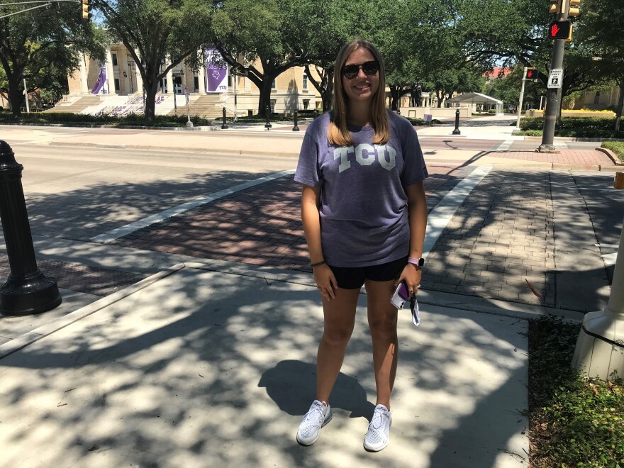 A photo of a young woman with blonde hair in a purple TCU shirt standing on a tree-shaded sidewalk.