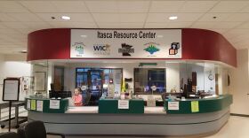 Two county employees sit behind glass at a large desk with a sign reading "Itasca Resource Center."
