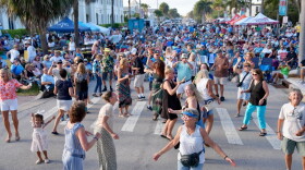 A large crowd fills a palm tree-lined street at an outdoor evening concert, with dozens of people dancing in the foreground and spectators seated in lawn chairs along the sidewalks under vendor tents in the background.