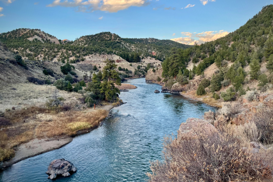 A river flows through a partially green, arid environment. There are higher inclines on either side of the river and what appears to be a railroad track running alongside it.