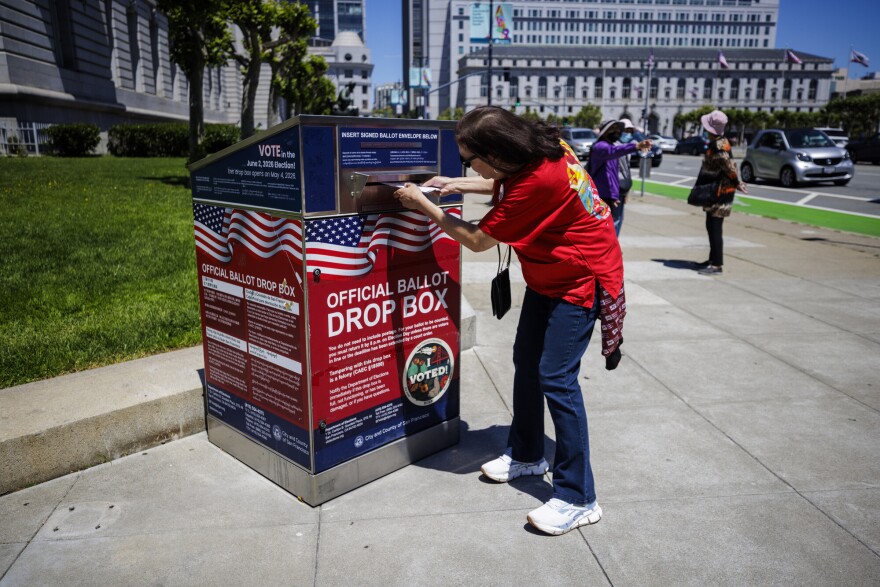 A woman wearing a red t-shirt and jeans leans down to see her ballot as she slips it into a red, white, and blue Official Ballot Drop Box on a city sidewalk. 