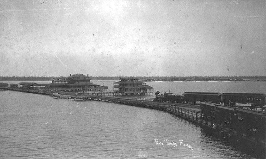 Photograph of Port Tampa, Florida, focusing on the pier, including railroad tracks. Two freight trains sit on the tracks, and three people stand on the pier.