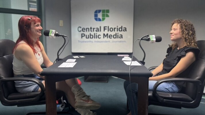Two young women sit across each other, microphones are in front of them, and they are smiling.