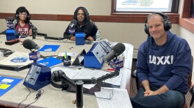 Three smiling people wearing headphones sit at a table in a radio talk studio. A woman at left has short black hair and is wearing a patterned back and white sweater and a maroon t-shirt; a man at center has shoulder-length black hair and is wearing a black sweatshirt; a man at right has short dark hair and is wearing a blue hoodie with white text that says "WXXI"