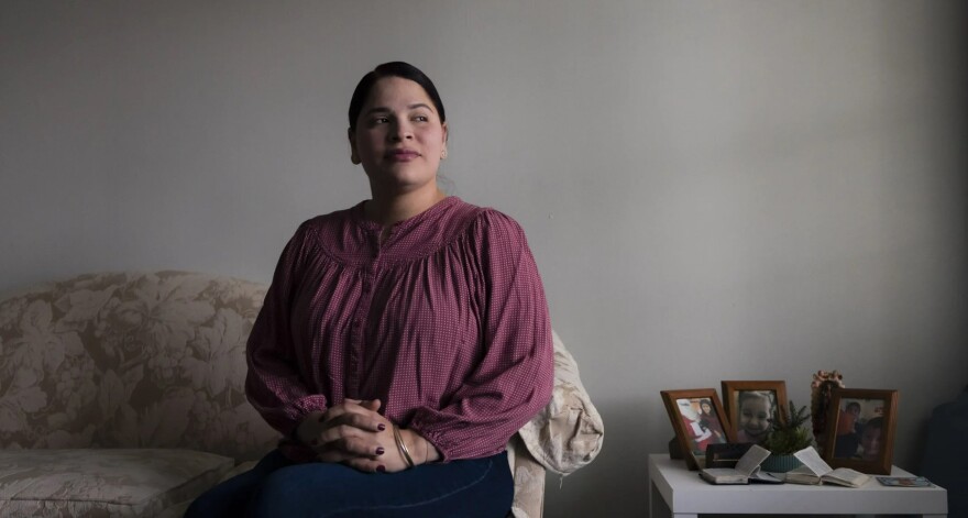 A woman with dark hair tied back wearing a blouse and jeans looks off into the distance while sitting in her living room.