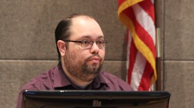A man with a scraggly beard and balding head at a desk behind a computer monitor