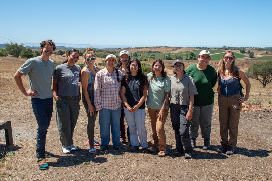 A group of people smile facing the camera outside in front of rolling hills on a sunny day. 