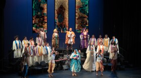 A stage filled with actors is in frame. A choir is on either side of the stage with three people standing in the middle slightly above them. There are three stained glass looking panels on the back wall.