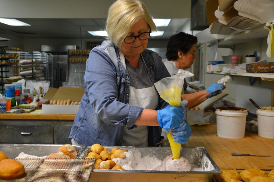  A woman wearing an apron fills a white powdered donut with yellow cream from a bag.