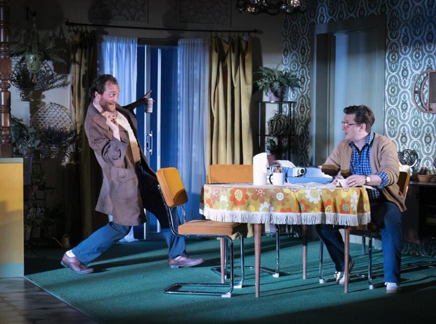 Man in coat stands by table where man sits