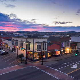 Shops and other buildings line an intersection in downtown Fort Bragg under a colorful evening sky.