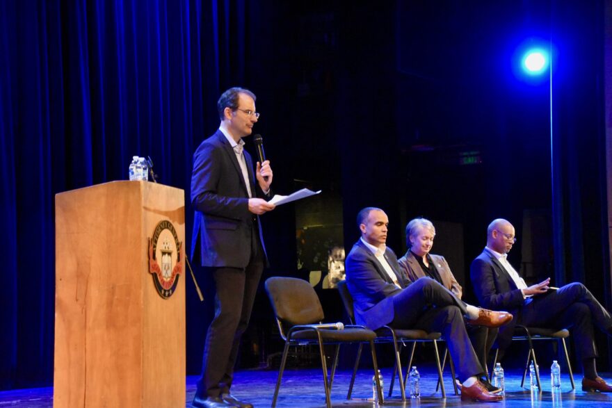 Colorado Attorney General Phil Weiser stands in a suit on stage with a microphone. Three other people sit in chairs on stage.