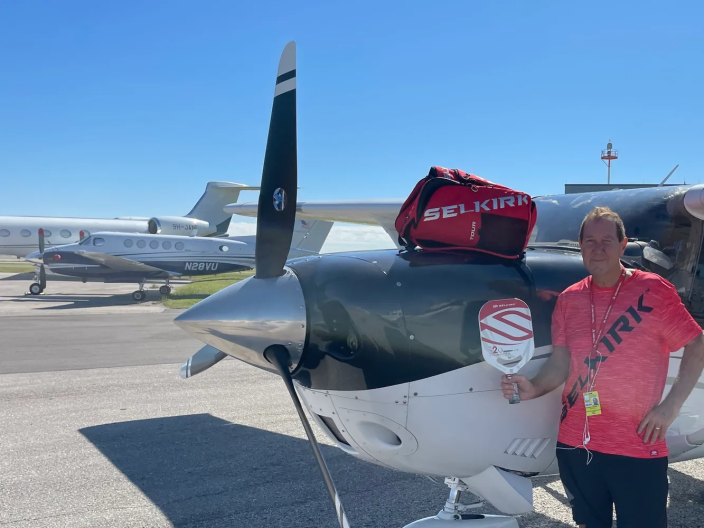 A man wearing a red shirt stands next to a plane with a Pickleball paddle in his hand. 
