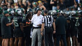A football coach talks to a referee while the team huddles behind them.