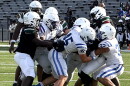 football action showing several players in a white uniform with blue strips tackling a player in a black jersey. both teams wearing white helmets. set on a green field with white football lines