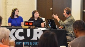 People are seated in an audience watching two women being interviewed by a man at a table. On the table sits an "on air" light and the table is covered by a table cloth that reads KCUR. The camera is positioned behind people in the crowd watching the interview. 