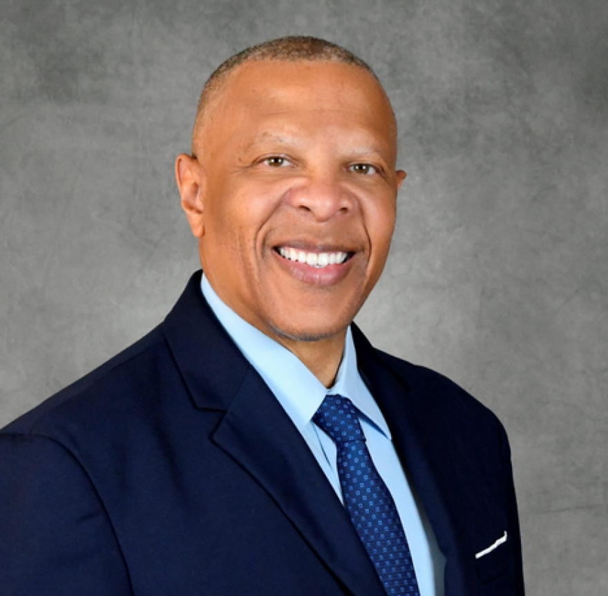 John Ewing is wearing a suit and tie and smiling against a gray backdrop