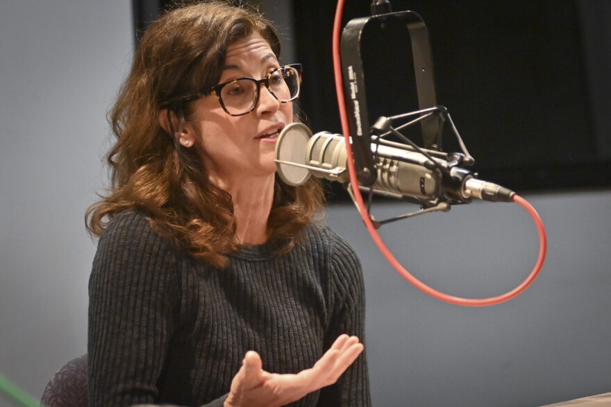 A woman sitting inside a radio studio talks at a microphone and gestures with her right hand.