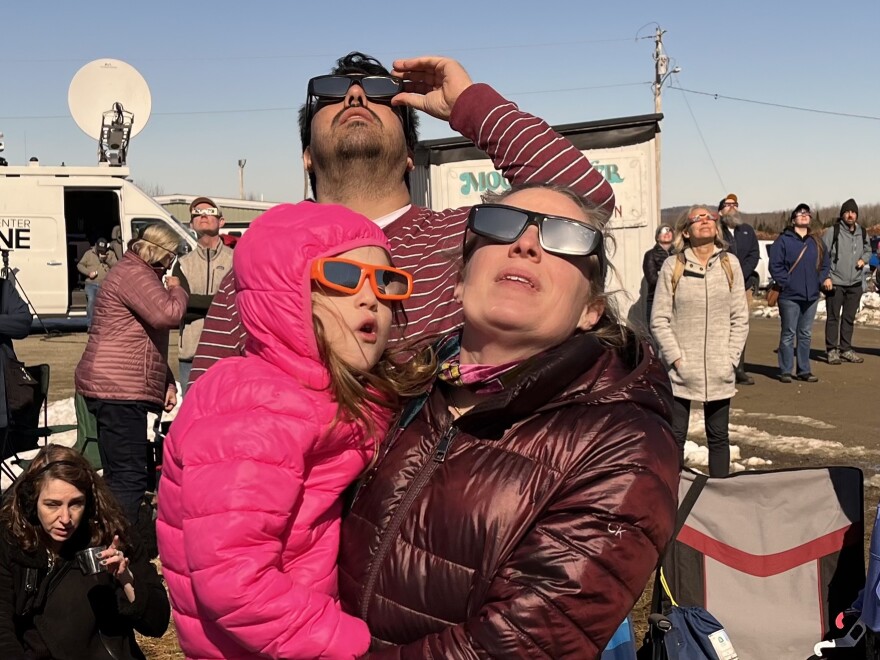Woman holding girl, both with eclipse classes gazing upward surrounded by many others doing the same