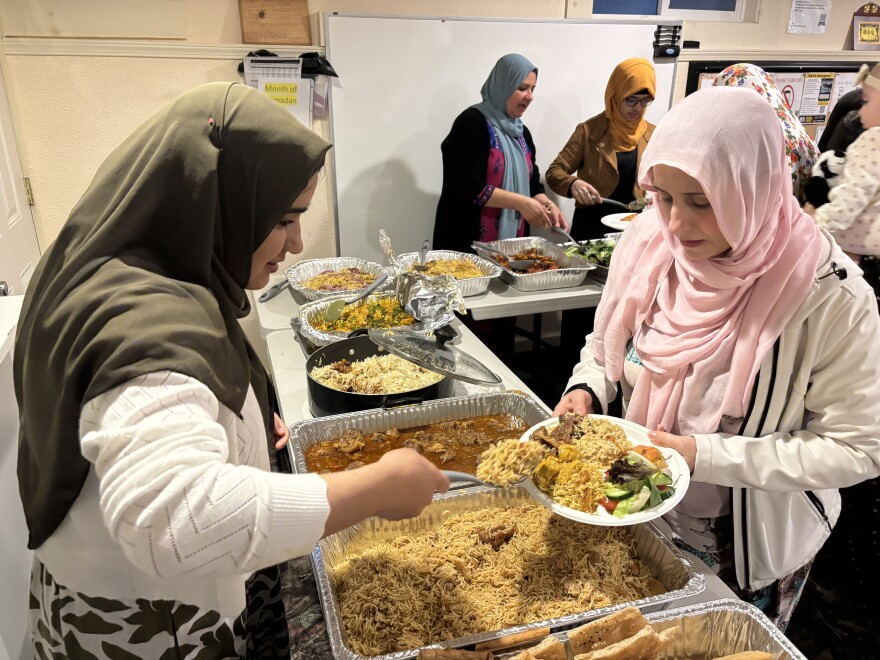 A woman is serving a dish of rice, chicken and lamb to another woman who is holding a plate. There are trays of food between them. The women on the left is wearing a green headscarf and the women on the right is wearing a pink headscarf.