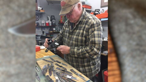 A man wearing a plaid patter shirt measuring silver knives with a table with a pile of knives in front of him.