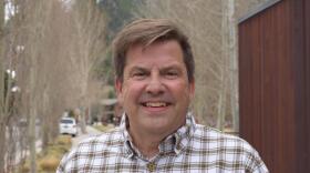 A man wearing a white and tan flannel shirt smiles for the camera while standing outside.