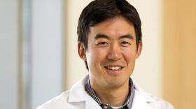 Sam Wang, a man with dark hair and wearing a white physicians jacket that reads "Children's Hospital Colorado" on one side and "G. Sam Wang, MD" on the other side smiles for a photo.