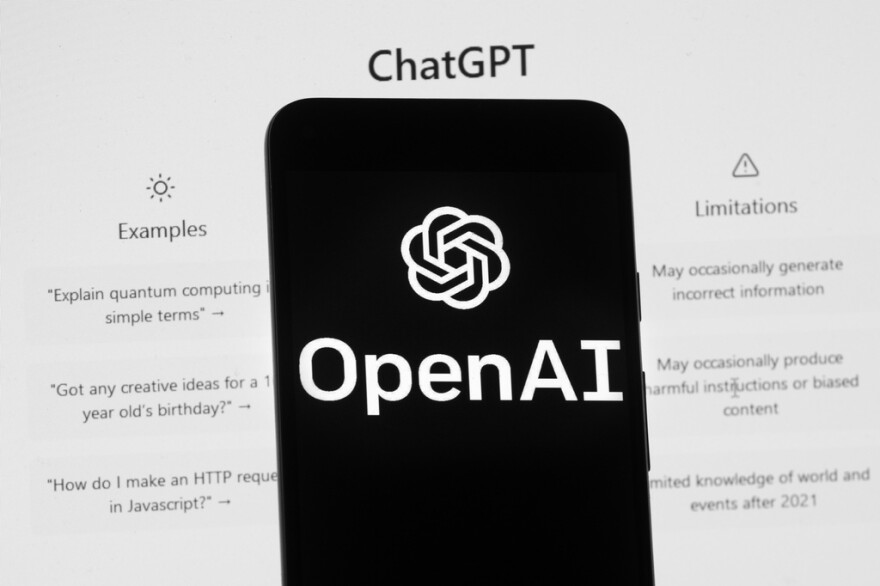 The OpenAI logo is seen on a mobile phone in front of a computer screen which displays the ChatGPT home screen.