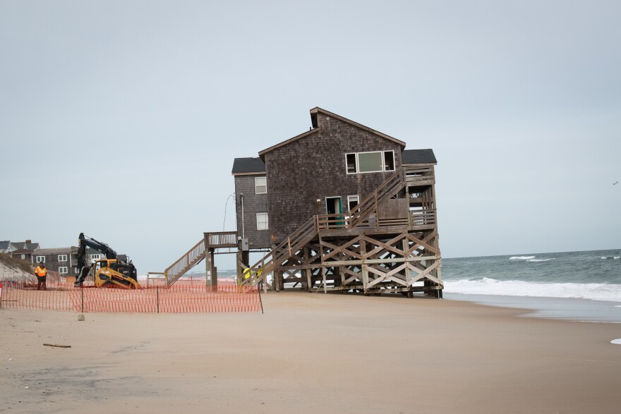 A construction vehicle prepares to demolish a house on stilts located very close to the shore.