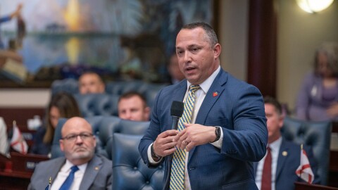 Man in suit and tie speaks into microphone with other representatives around him 