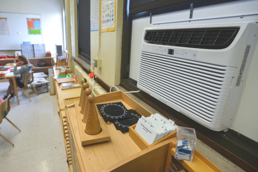 Interior photo of a school classroom that shows one out-of-focus student sitting far left. At right, in foreground, is a 