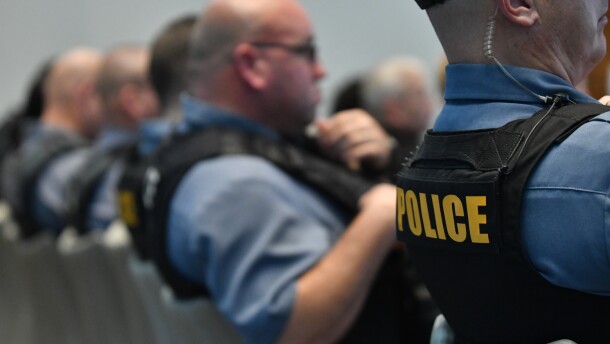 Four police officers sit in a row of chairs, backs to the camera. In the foreground, the back of a protective vest reads, "police."