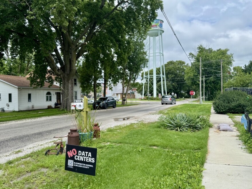 A yard sign saying "no data centers in Bloomington-Normal and McLean County" sits in a grassy easement next to a mailbox with antique milkjugs around it.