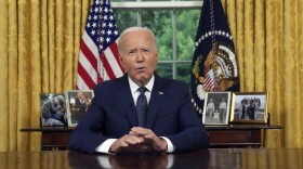 Man in blue suit and white shirt sitting at the Resolute Desk in the White House Oval Office. He is looking at the camera and speaking.