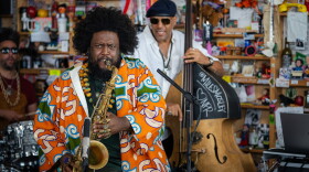 A Black man plays saxophone in a colorful jacket with several rings on each finger. Behind him are two people playing an upright base and the drums