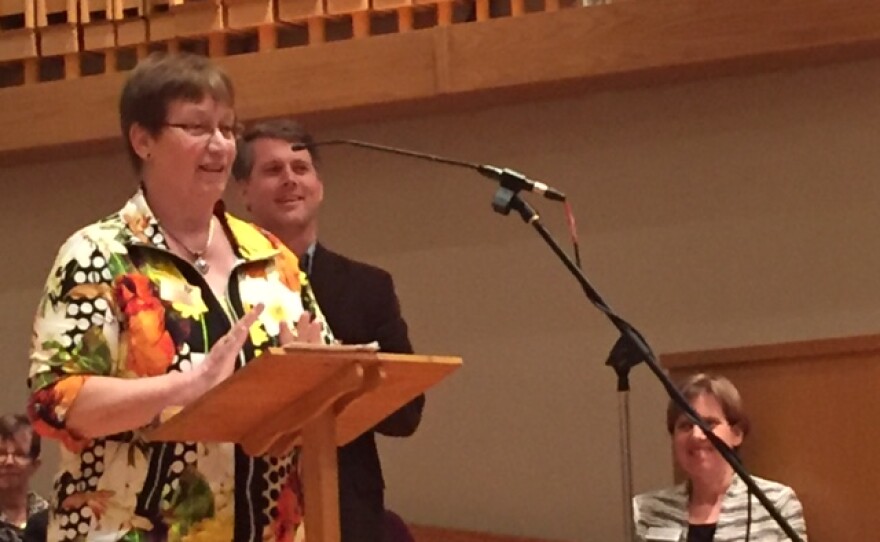 Woman at podium with back in background and woman sitting in a church pew stage left