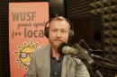 Man in suit jacket and button down shirt sits behind microphone with headphones on. A pink WUSF Your Spot for Local is behind him. 