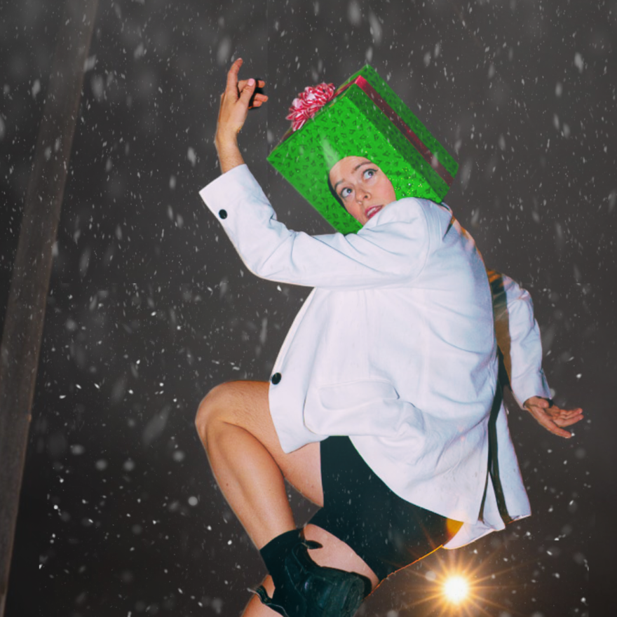 Holiday music, dancing Santas and present-box heads are all in store during Bare Bait Dance’s A Few of My Favorite Things holiday production. The photo shows a performer wearing a green gift box on their head, dressed in a white blazer and black boots, striking a dynamic dance pose amid falling snow. 