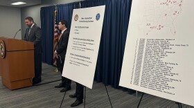 Men in suits stand at a podium next to a large easel with a map on it. The map shows 29 NCAA basketball games that allegedly involved point-fixing, including a February 24, 2024 game between Western Michigan University and The State University of New York at Buffalo. According to prosecutors, players on Buffalo's team engaged in points-fixing during the game.