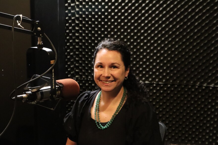 A woman sits smiling in a recording studio beside a microphone.