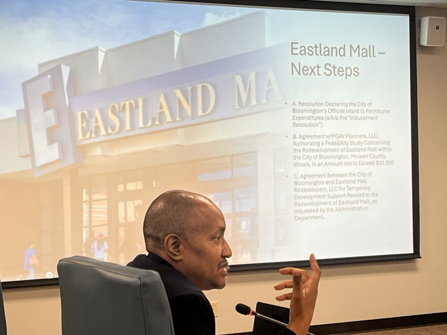 In the foreground, a man addresses someone offscreen. A slideshow is displayed behind him 